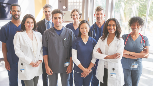 Multiple medical professionals in scrubs, doctor's coats, and dress clothes are all smiling in a group.